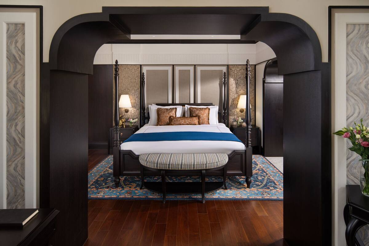 A four-poster bed with a blue and white bedspread, flanked by two nightstands with lamps, set against a dark wooden floor and a patterned rug.