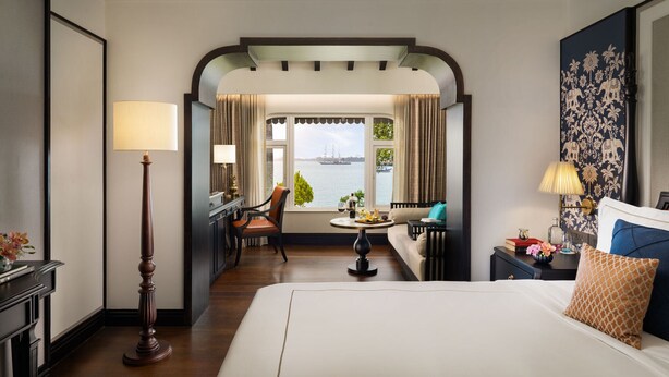 Una habitación de hotel con una cama grande, un escritorio con lámpara, una silla, una mesita con lámpara y vista al agua a través de una ventana.