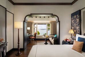 A hotel room with a large bed, a desk with a lamp, a chair, a small table with a lamp, and a view of the water through a window. - Taj Malabar Resort & Spa, Cochin (Kochi)