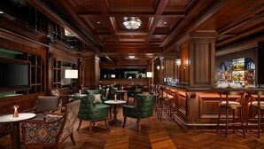 Zona de bar con paneles de madera, sillas tapizadas en verde y una barra con una amplia selección de licores.