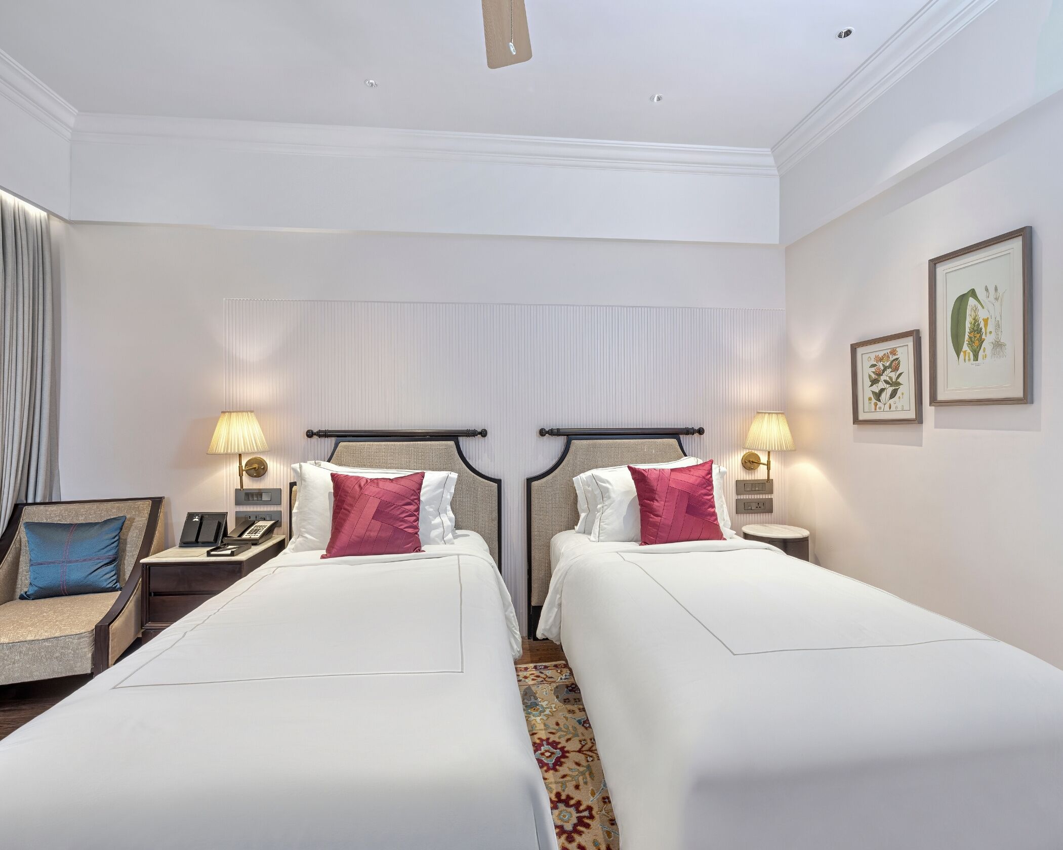 A hotel room with two beds, a desk, a chair, a telephone, and wall-mounted lamps.