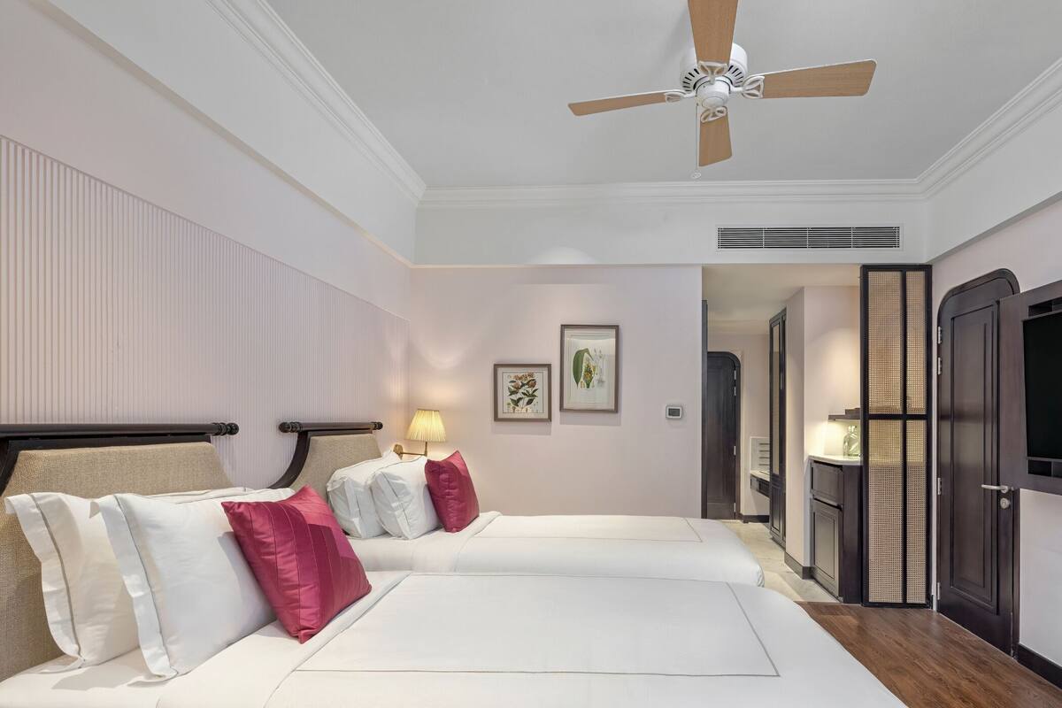 A hotel room with two beds, a ceiling fan, a wall-mounted lamp, and a door leading to a bathroom.