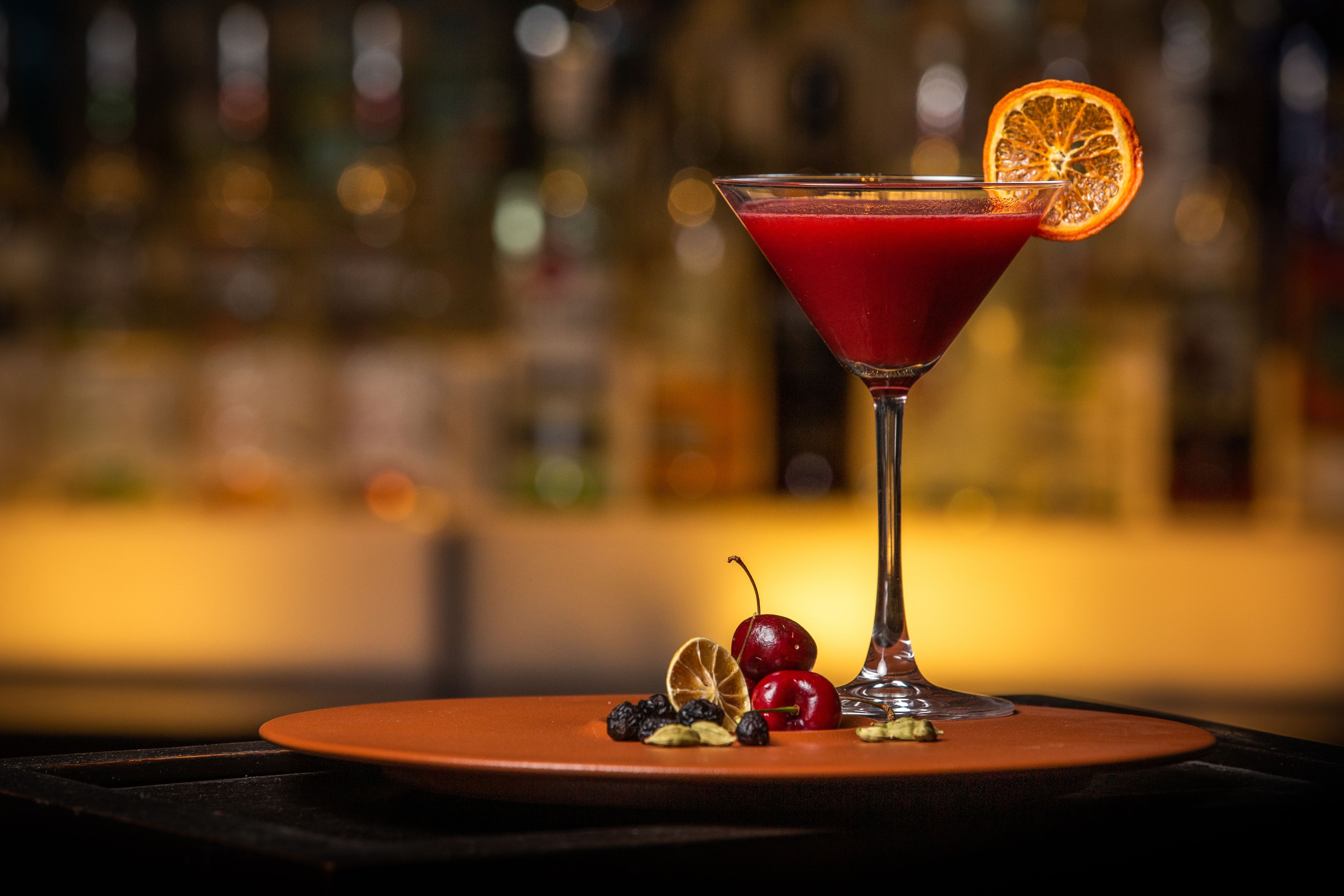 Una copa de martini con una bebida roja, adornada con una rodaja de naranja y una cereza, sobre una mesa con un fondo desenfocado de botellas.