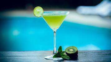 Un vaso de cóctel con una rodaja de lima, un trozo de kiwi y hojas de menta sobre una mesa junto a una piscina.