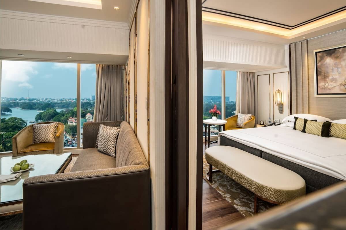 Une chambre d’hôtel avec un grand lit, un canapé, une petite table ornée d’un vase de fleurs, et une vue sur un paysage urbain et une étendue d’eau.