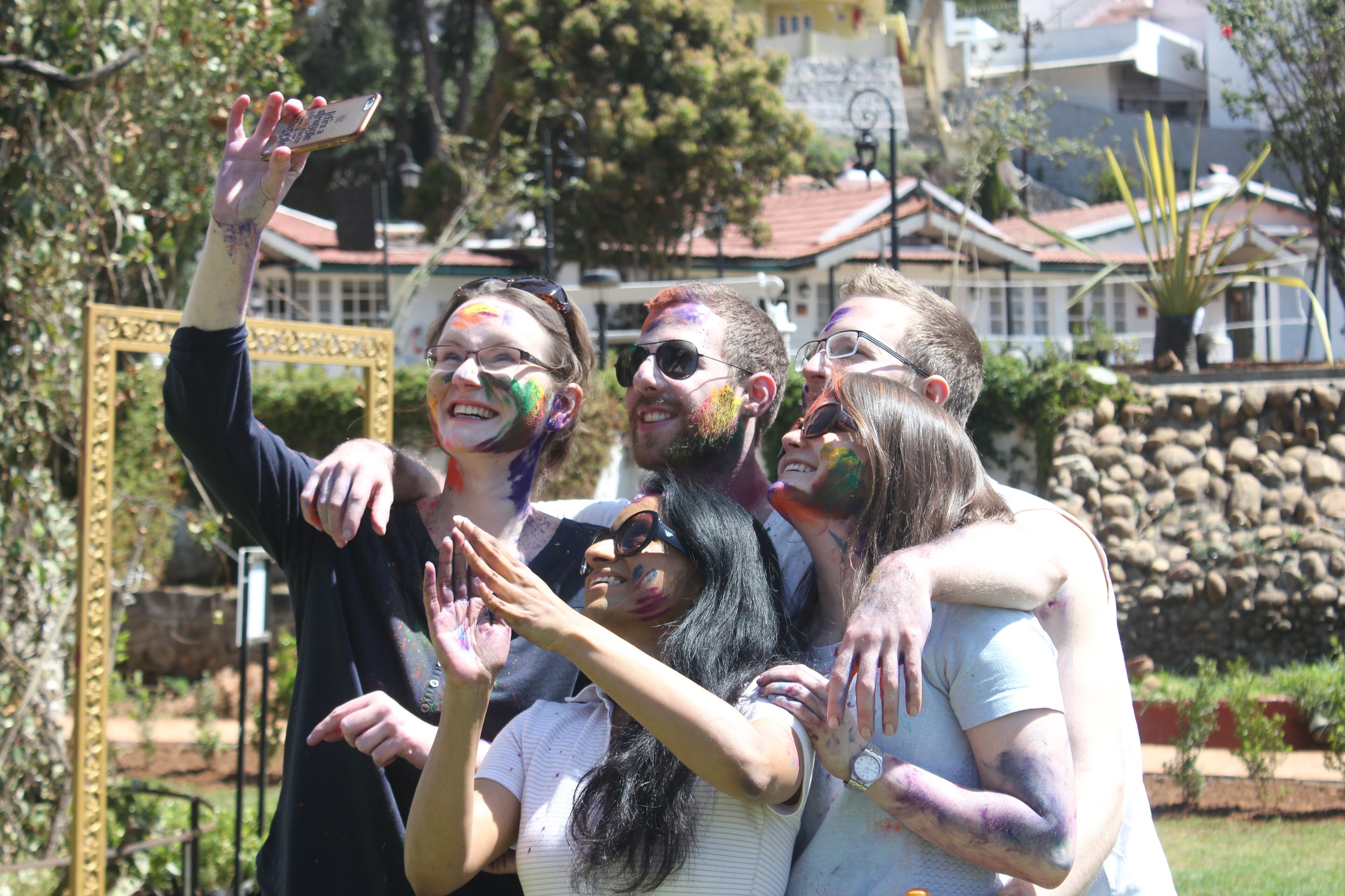 A group of people with colorful paint on their faces and arms, standing in front of a building with a stone wall and a sign.