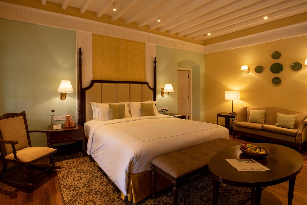A hotel room with a large bed, a wooden chair, a small table with a bowl of fruit, and a sofa.