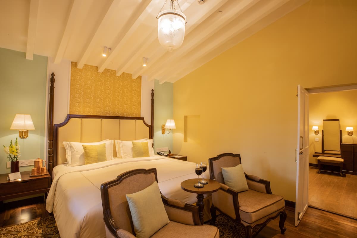 A hotel room with a large bed, two armchairs, a small table with a wine glass, and a chandelier.