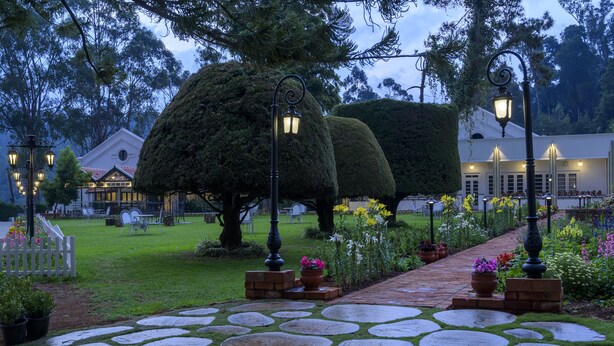 Un sendero de piedra conduce a un edificio de fachada blanca y techo rojo, rodeado de setos cuidados y plantas en flor.