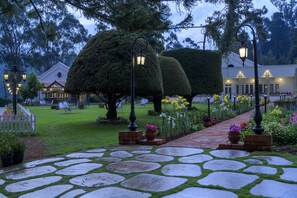 Un sendero de piedra conduce a un edificio de fachada blanca y techo rojo, rodeado de setos cuidados y plantas en flor.