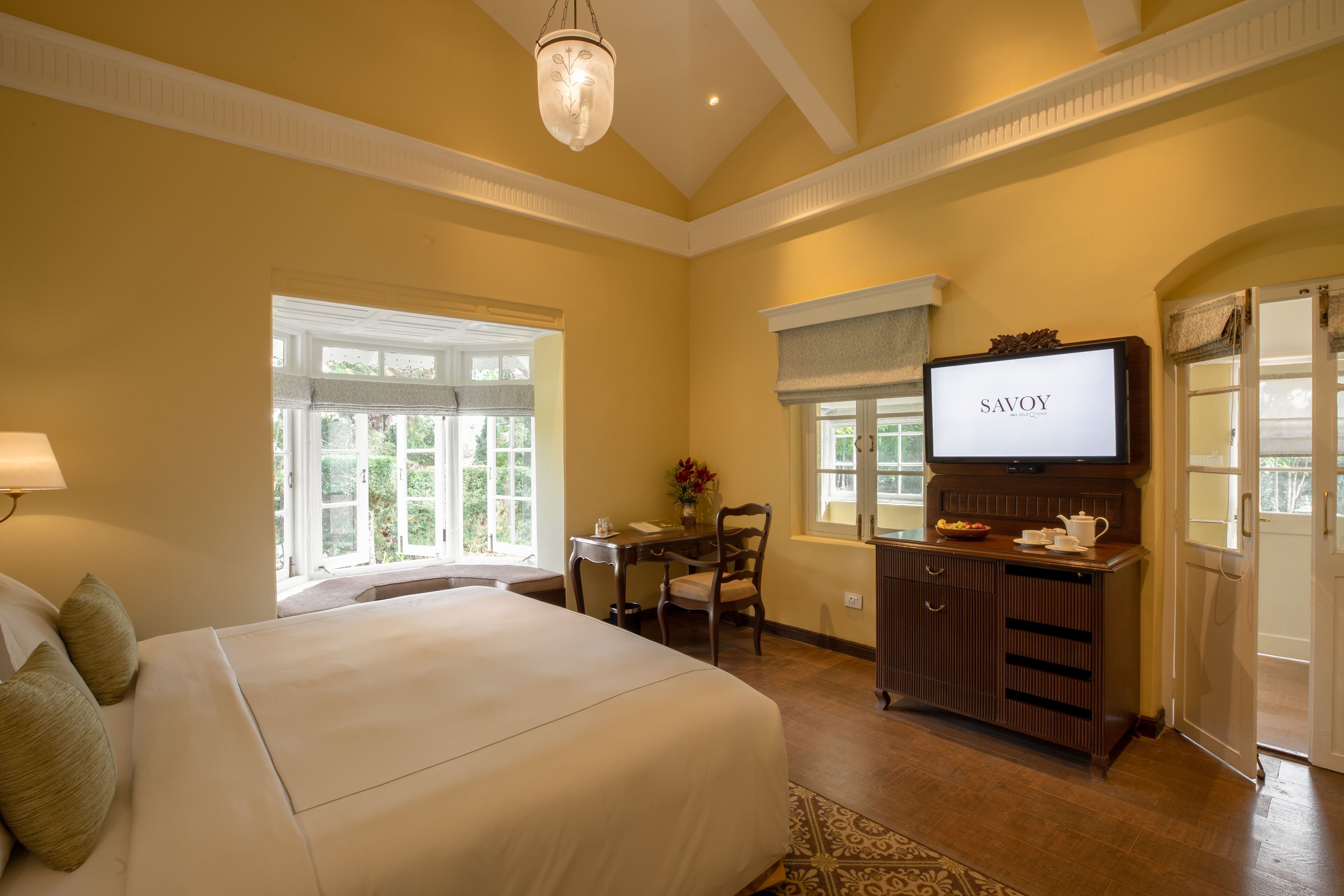 A hotel room with a large bed, a desk with a television, and a view of greenery outside.