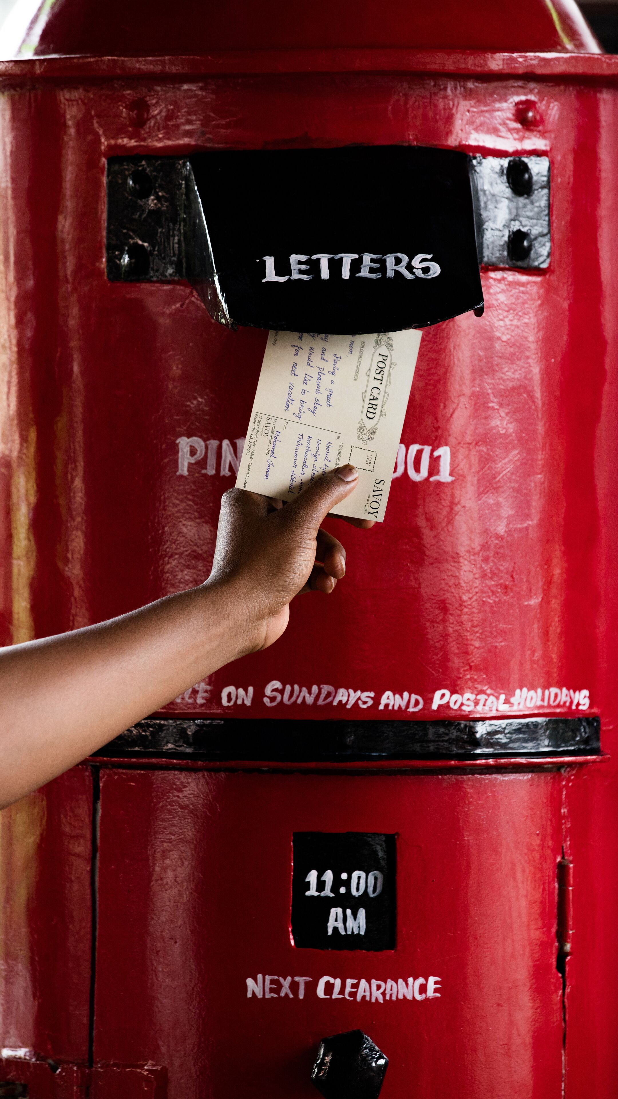 Uma mão depositando uma carta em uma caixa de correio vermelha com a etiqueta 'Letters', um aviso de 'Próximo Despacho' e um horário fixado indicando '11:00'. 