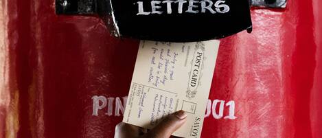 A hand placing a letter into a red mailbox labeled "Letters" with a "Next Clearance" sign and a posted schedule of "11:00 AM" and "Next Clearance".