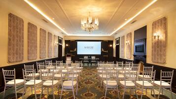 Une salle de conférence dotée d’un grand écran affichant le mot « SAVOY », de rangées de chaises blanches, d’un lustre, d’un papier peint à motifs et d’un sol recouvert de moquette.