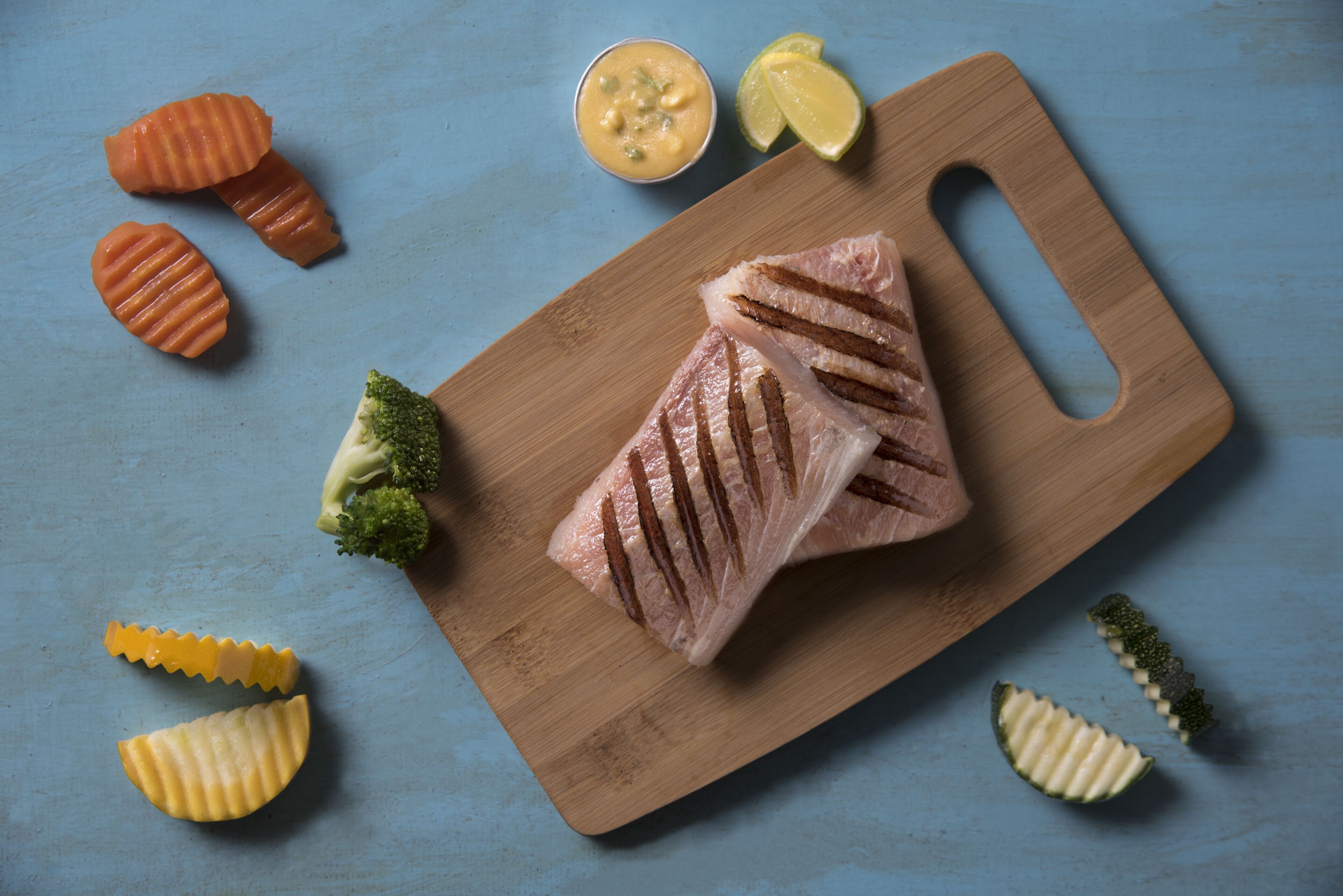 Una tabla de madera con un trozo de carne y diferentes verduras sobre una superficie azul.