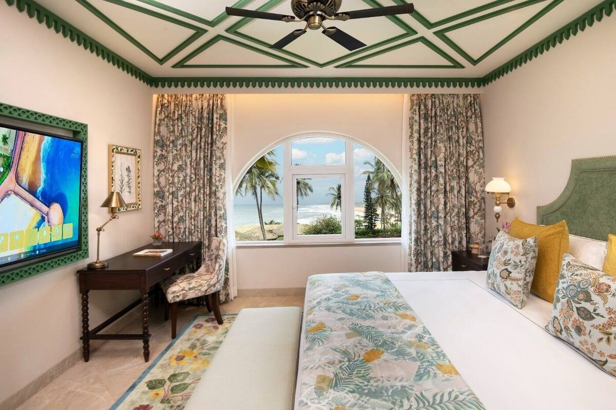 A hotel room with a large bed, a desk, a chair, and a TV. There is a window with a view of palm trees and the beach.
