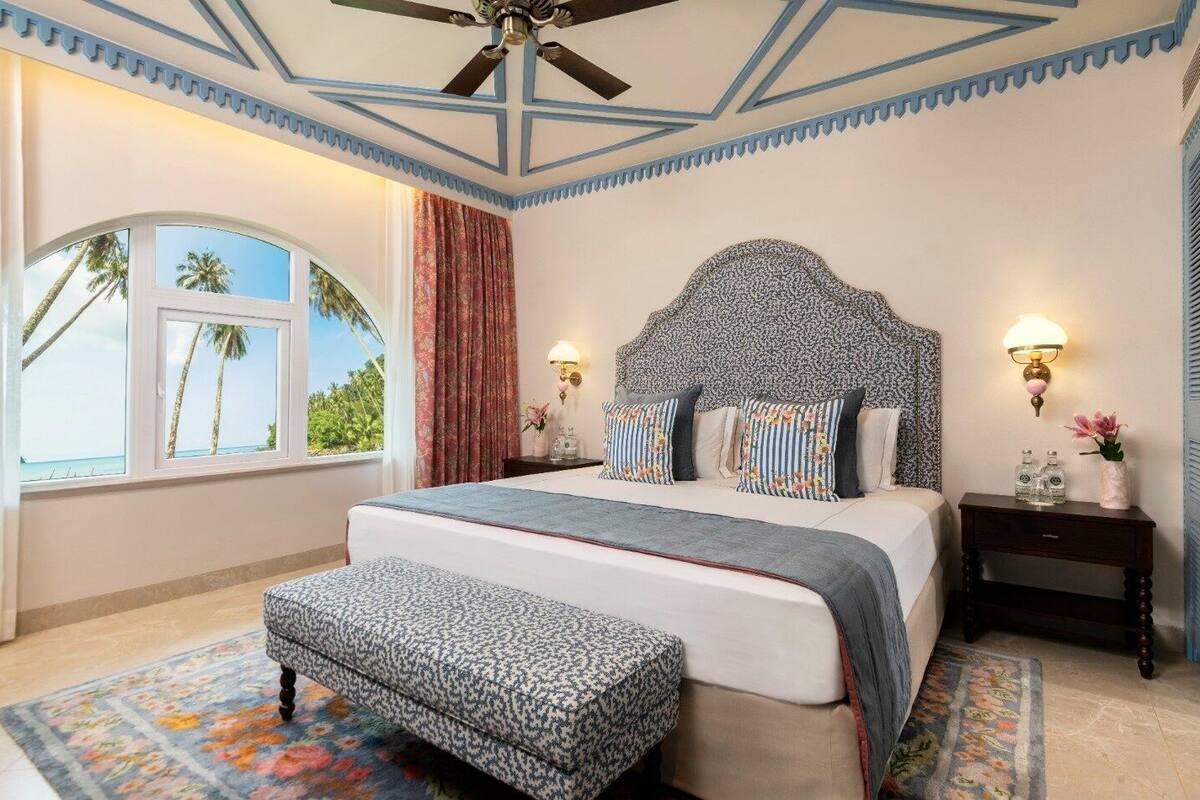 A bedroom with a large bed, a ceiling fan, a nightstand with a lamp, and a window with a view of palm trees.