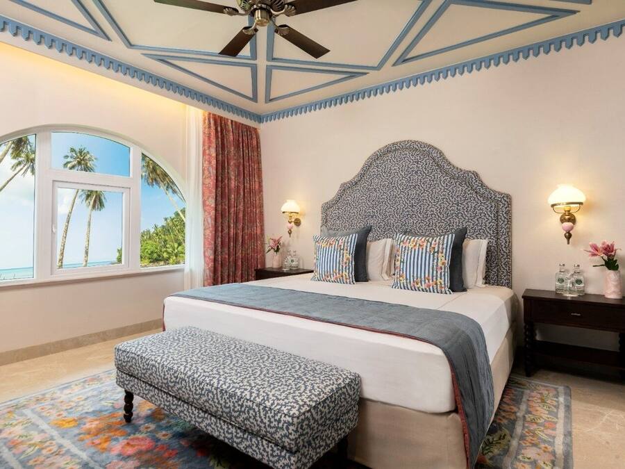 A bedroom with a large bed, a ceiling fan, a nightstand with a lamp, and a window with a view of palm trees.
