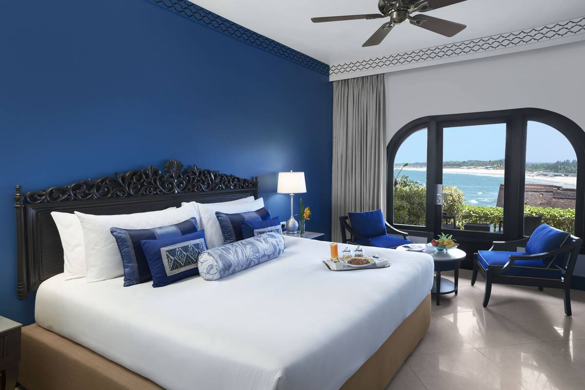 A hotel room with a large bed, a view of the beach, a ceiling fan, and a small table with breakfast items.