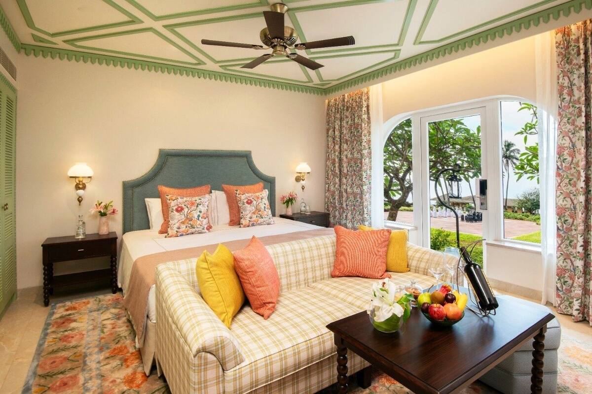 A bedroom with a bed, a sofa, a nightstand, a table with fruit, and a window with curtains.