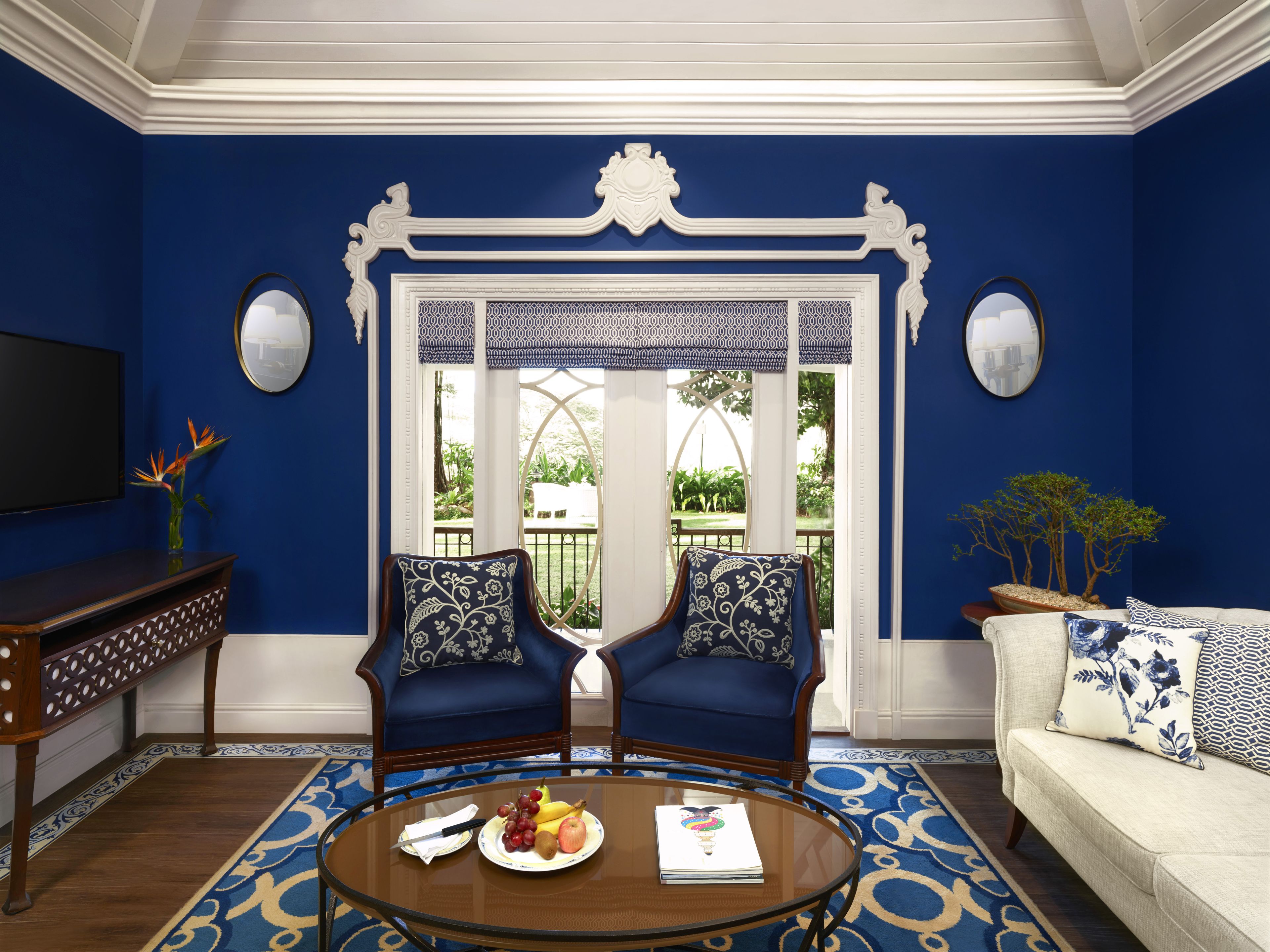 A room with blue walls, a white ornate doorway, two blue armchairs with patterned cushions, a round glass coffee table with fruits and a notebook, and a white sofa with patterned pillows.