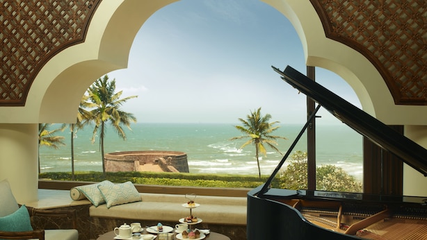 A room with a piano, a sofa, a table with tea and pastries, and a view of the ocean and palm trees. - Taj Fort Aguada Resort & Spa, Goa (Candolim)