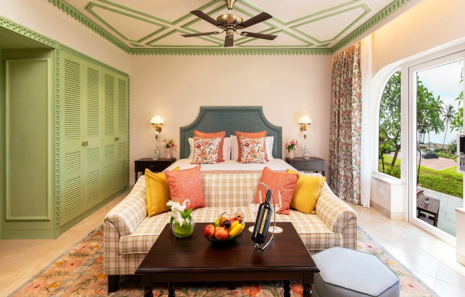 A bedroom with a bed, a sofa, a coffee table with fruit, and a view of the outdoors.