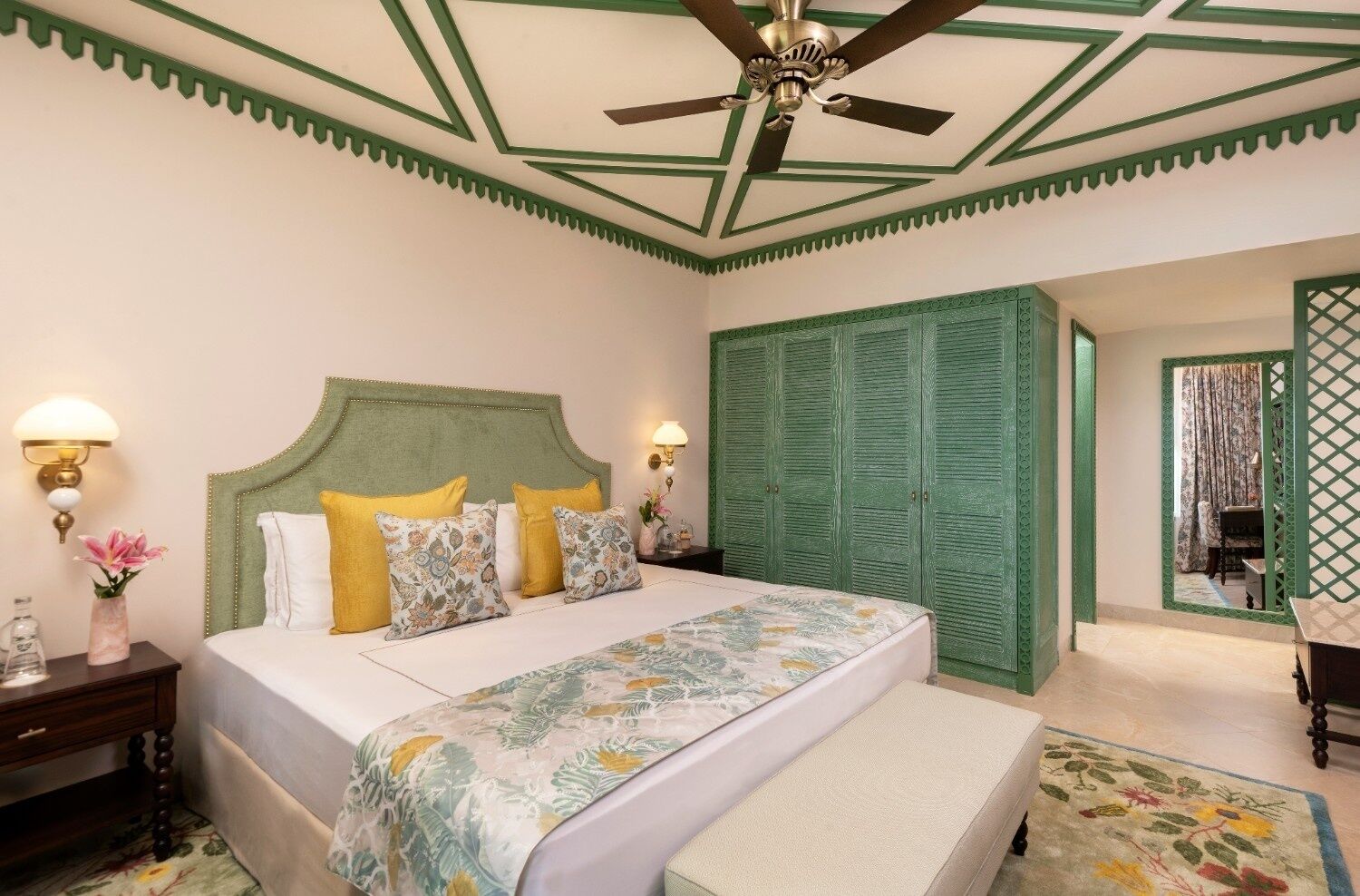 A bedroom with a bed, bedside tables, a nightstand, a green wardrobe, a ceiling fan, and a mirror.