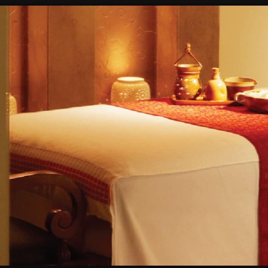 A neatly made bed with a white bedspread and a red blanket. There is a small table with a bowl of fruit and a bottle.