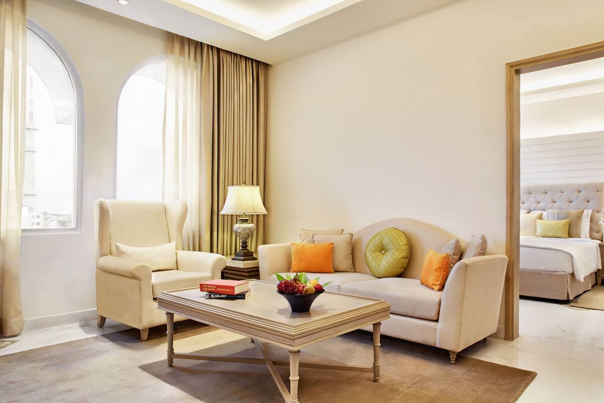 A hotel room with a sofa, armchair, and a bed with a headboard. There is a table with a lamp and a bowl of fruit.
