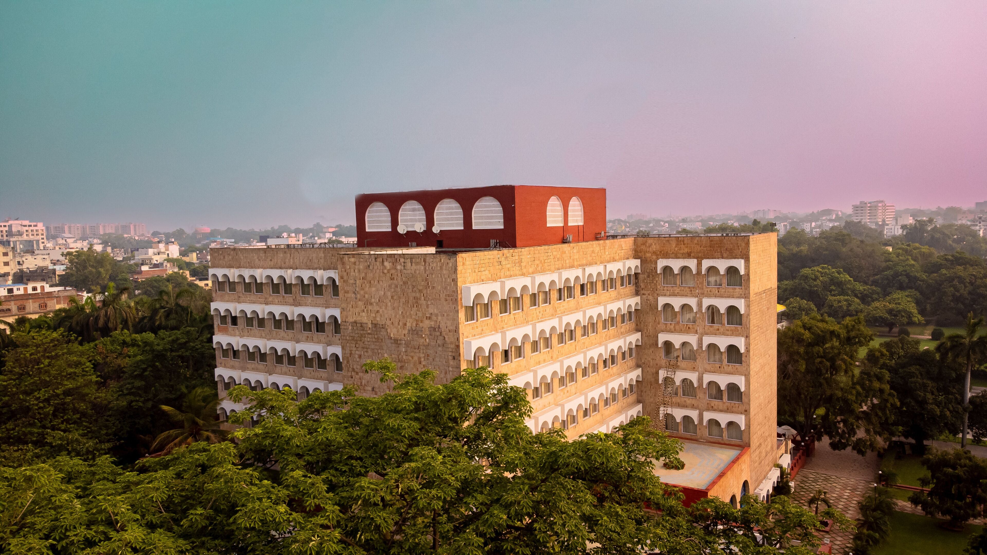 A multi-story building with a distinctive red section on top, surrounded by lush greenery.