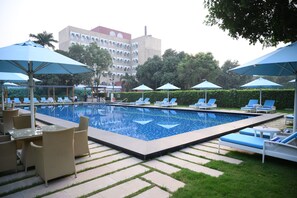 A swimming pool with lounge chairs and umbrellas, surrounded by a hedge and a building in the background.