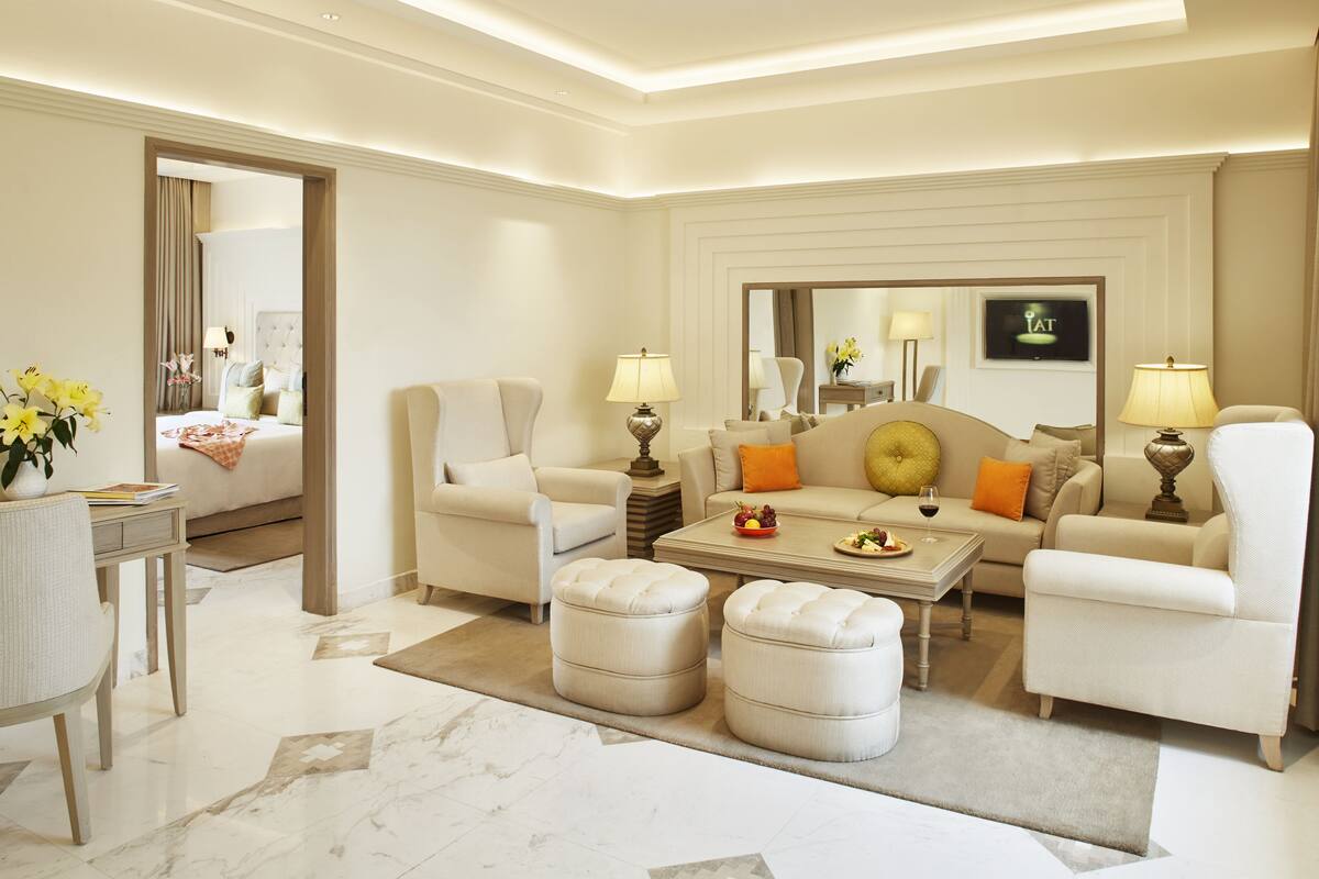 A modern hotel room with a living area featuring a sofa set, ottomans, a coffee table, and two lamps. A bed with a headboard is visible in the background.
