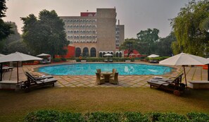 A swimming pool with lounge chairs and umbrellas, surrounded by greenery and a building with arched windows.