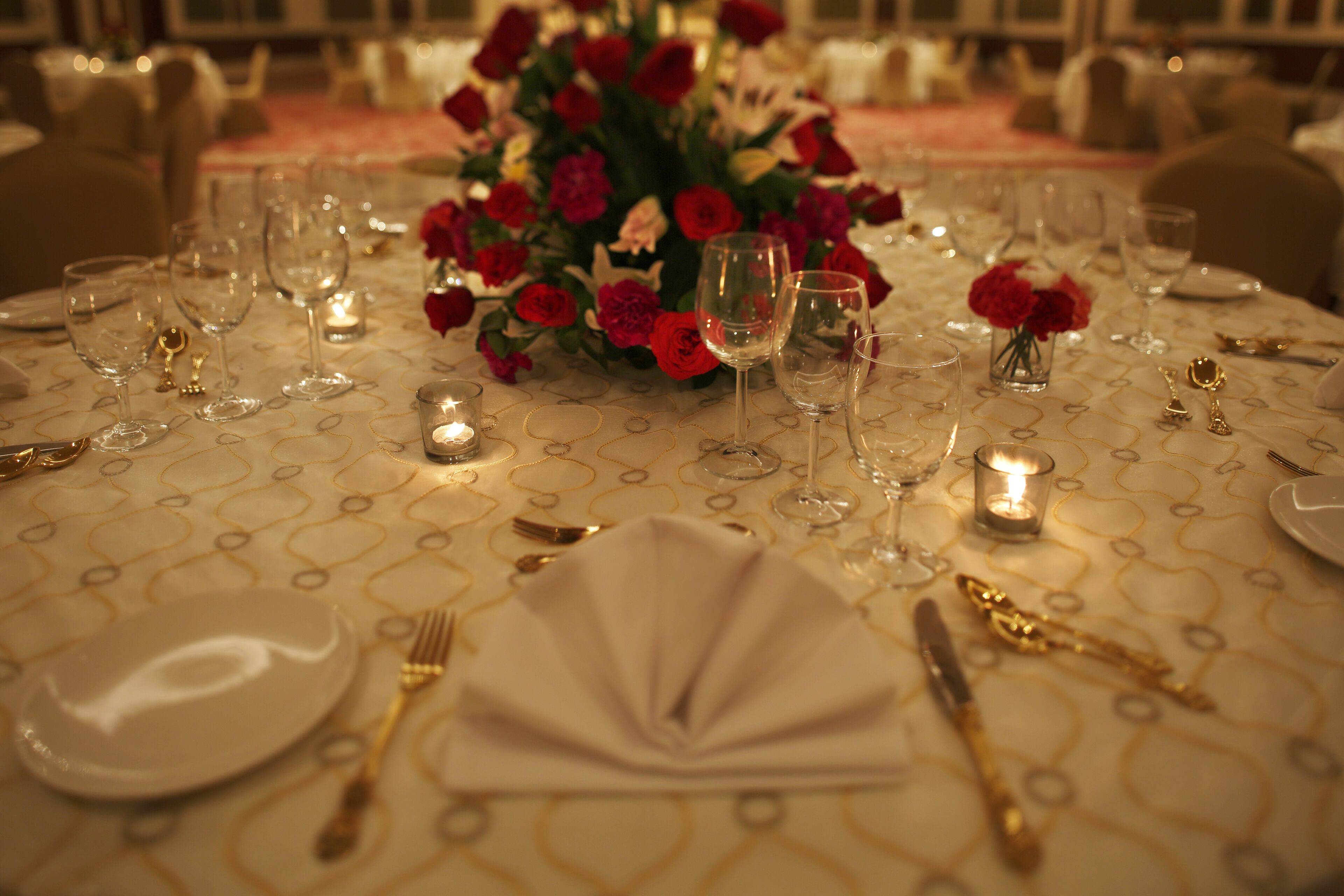 Une table ronde dressée pour un repas formel, avec une nappe blanche, des couverts dorés et des roses rouges dans un vase.