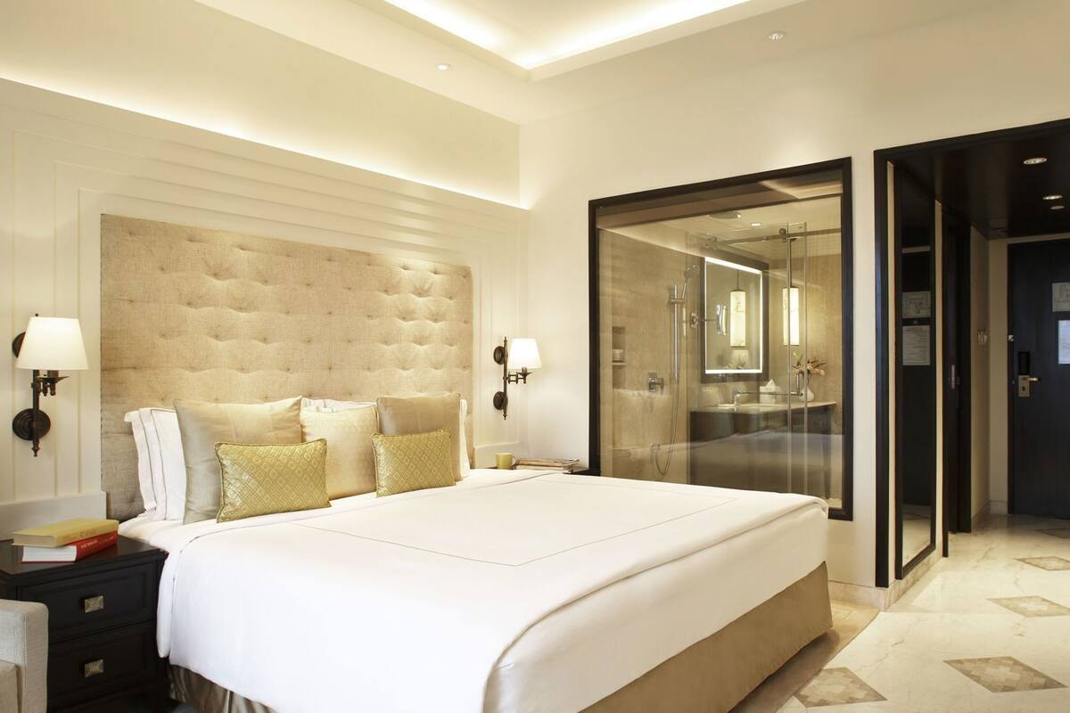 A modern hotel bedroom with a large bed, bedside tables, a nightstand, a wall-mounted lamp, and a glass-enclosed shower area.