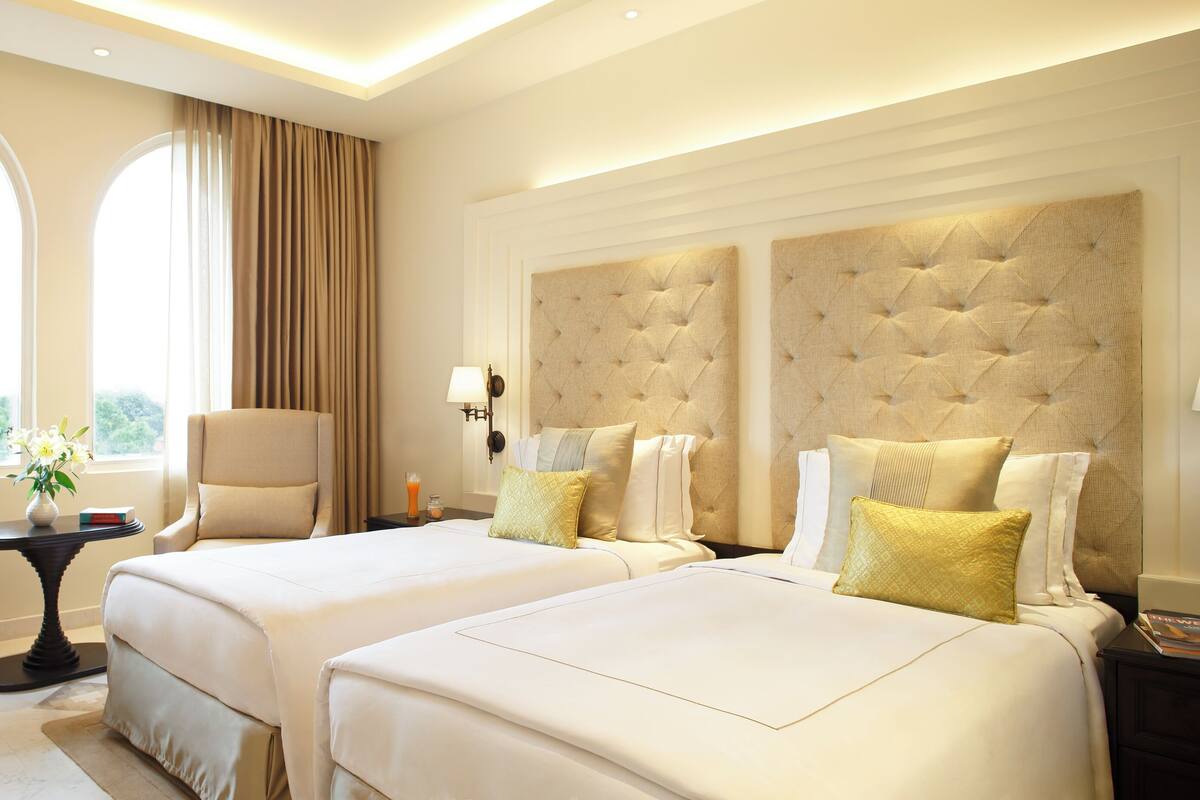 A hotel room with two beds, a TV, a small table with a vase of flowers, and a window with curtains.
