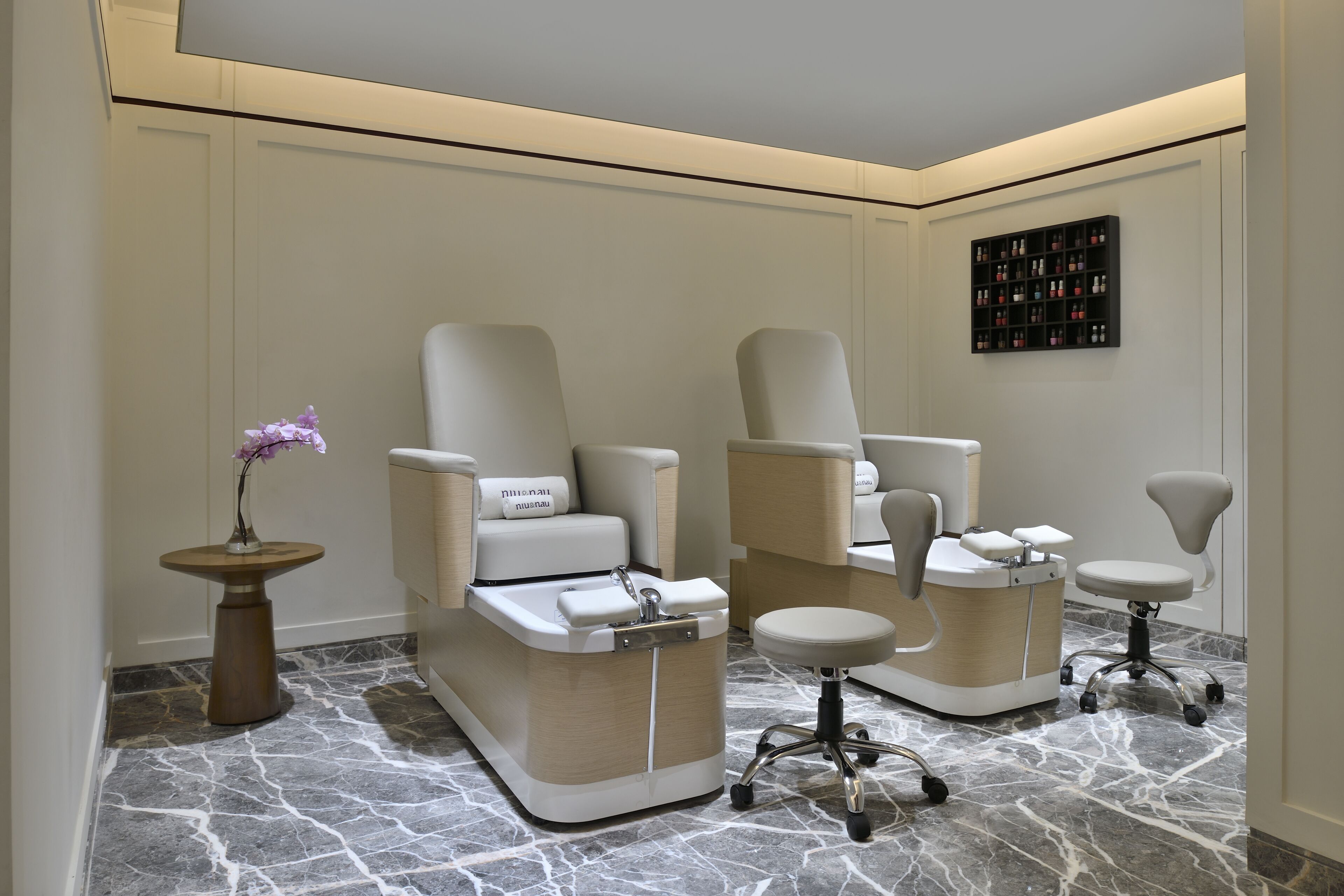 A spa room with two massage chairs, a small table with a vase of flowers, and a shelf with bottles.