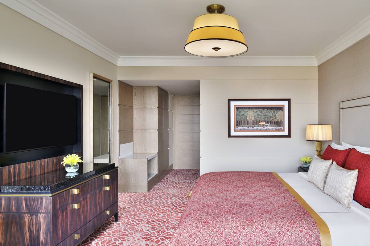 Habitación de hotel con una televisión de pantalla plana grande, un aparador de madera, una alfombra roja estampada, un sofá blanco con cojines rojos y blancos, y un cuadro en la pared.