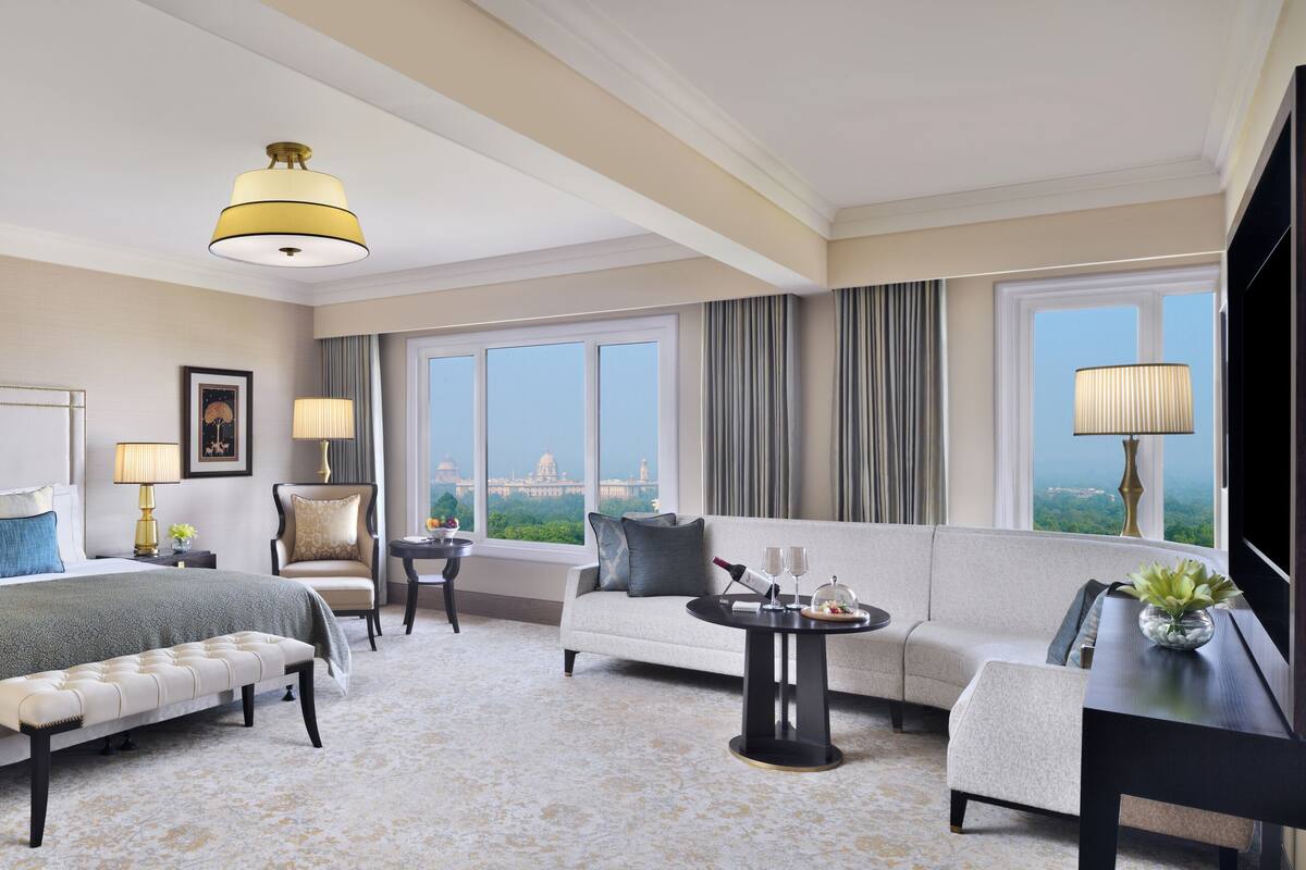 Habitación de hotel con una cama grande, un sofá, un banco, una mesita y vistas a un paisaje urbano a través de las ventanas.