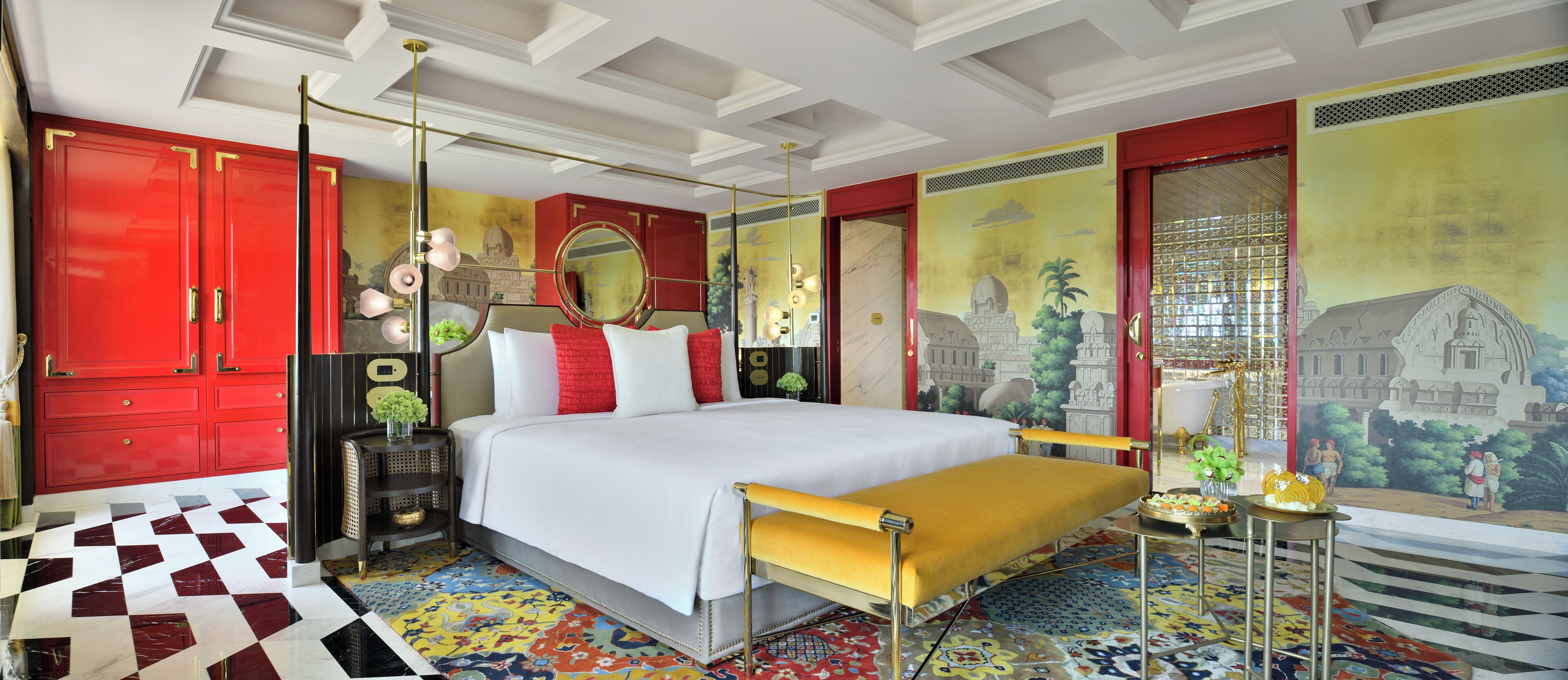 Une chambre d’hôtel avec un grand lit, une armoire rouge, un banc jaune et un sol aux motifs colorés.