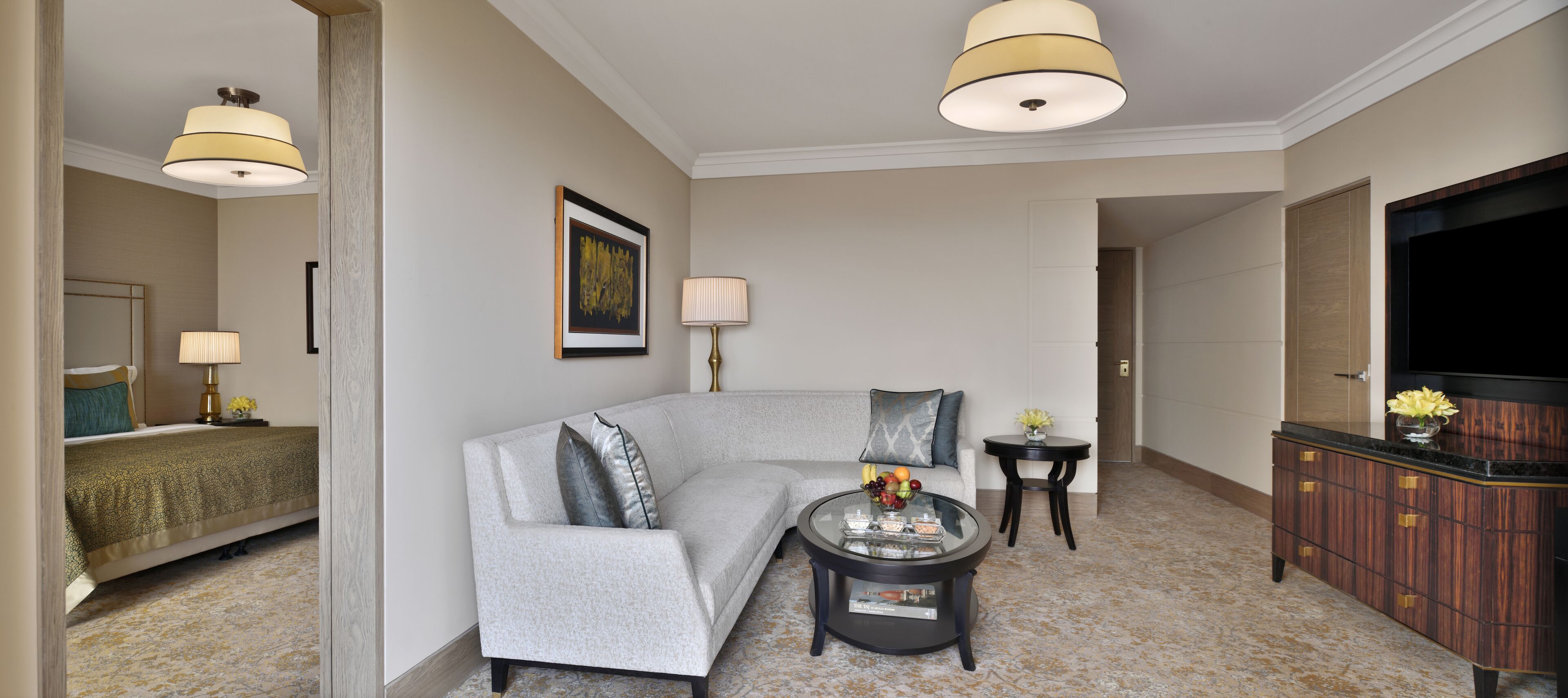 Quarto de hotel moderno com sofá cinzento, uma mesa de centro redonda com frutas, uma televisão, uma cama com colcha verde e uma pequena mesa com abajur.