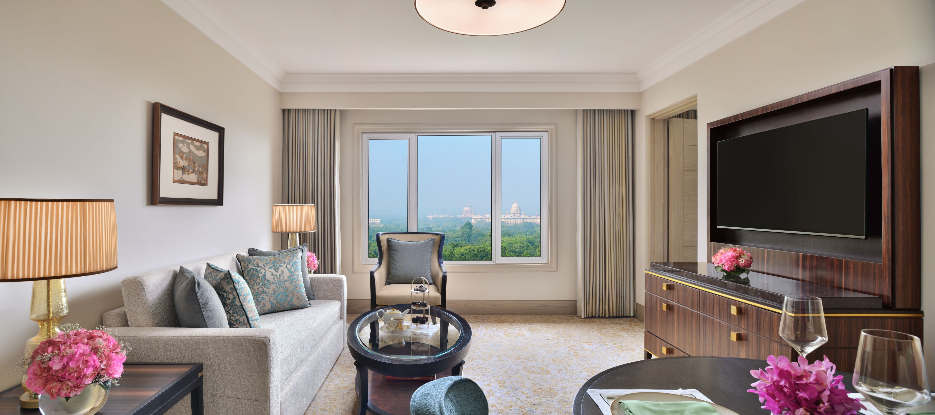 A hotel room with a sofa, armchair, coffee table, TV, and a view of a cityscape.
