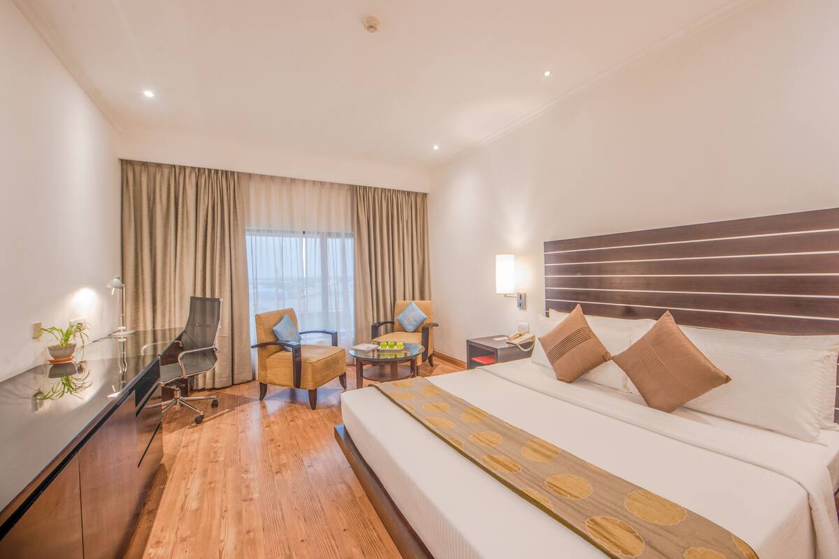 A hotel room with a large bed, a desk with a lamp, two armchairs, a small table with a plant, and a window with curtains.