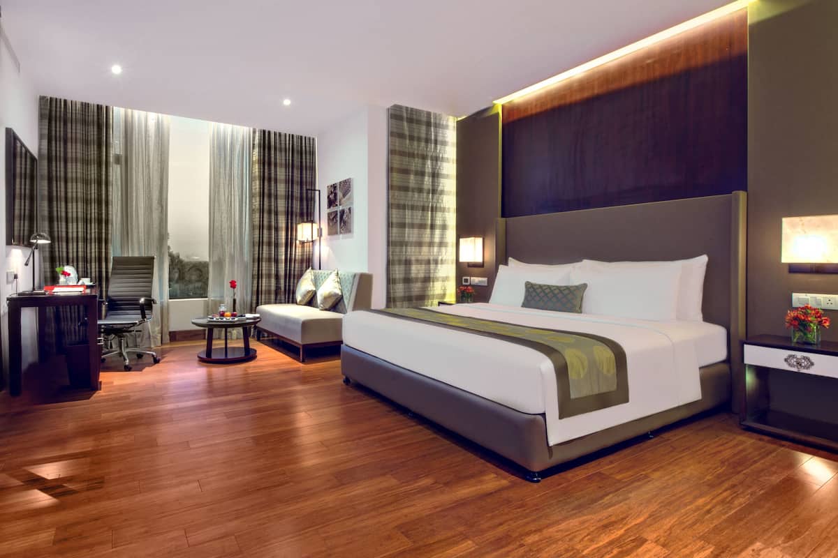 A modern hotel room with a large bed, a desk, a sofa, and a view of the cityscape.