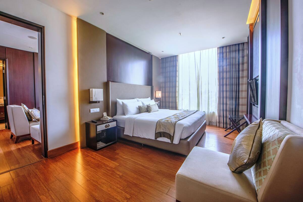 A modern hotel room with a large bed, a sitting area with a sofa, a desk with a lamp, and a view of the city through large windows.