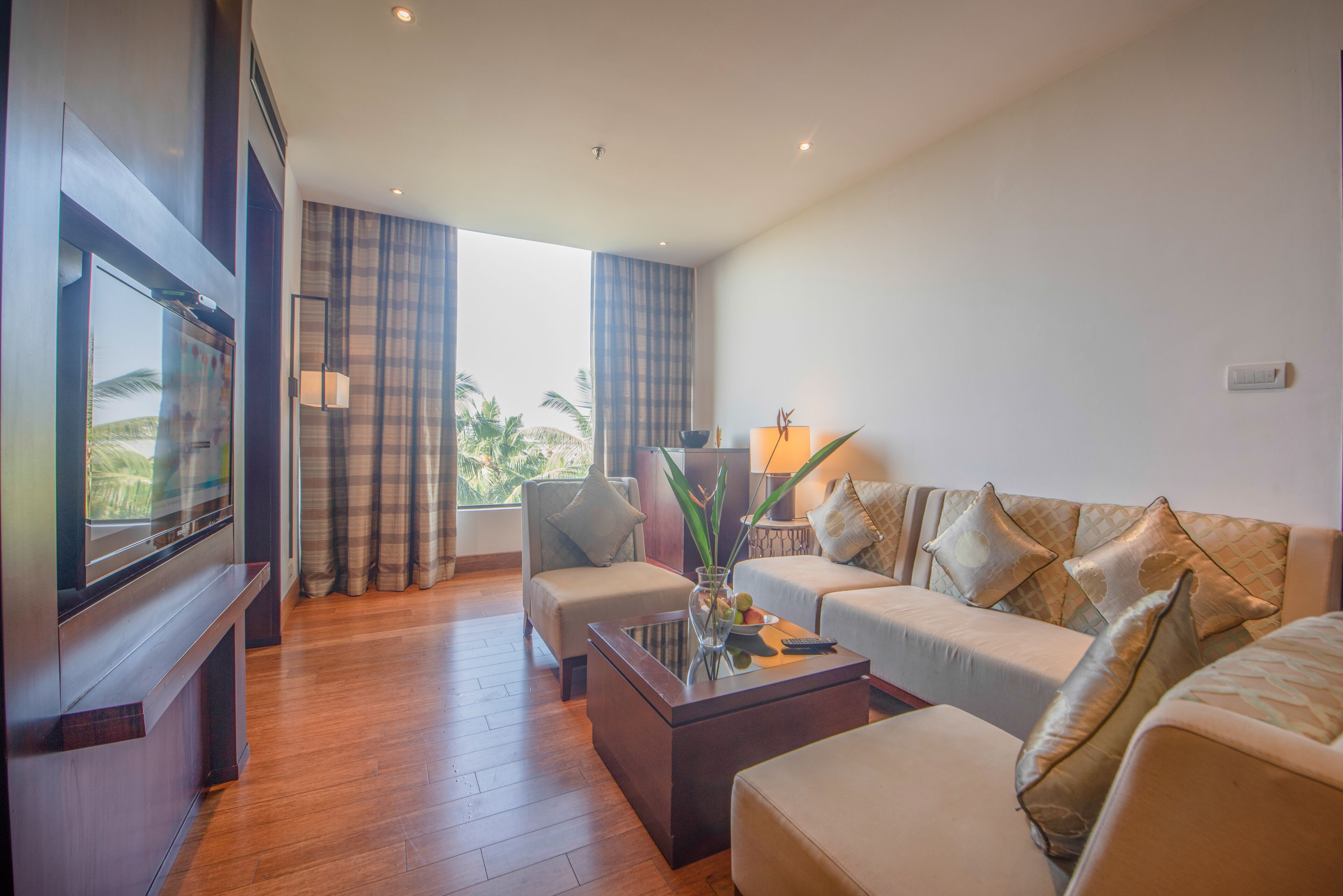 A modern living room with a flat-screen TV, a sofa, a coffee table, and a view of palm trees through large windows.