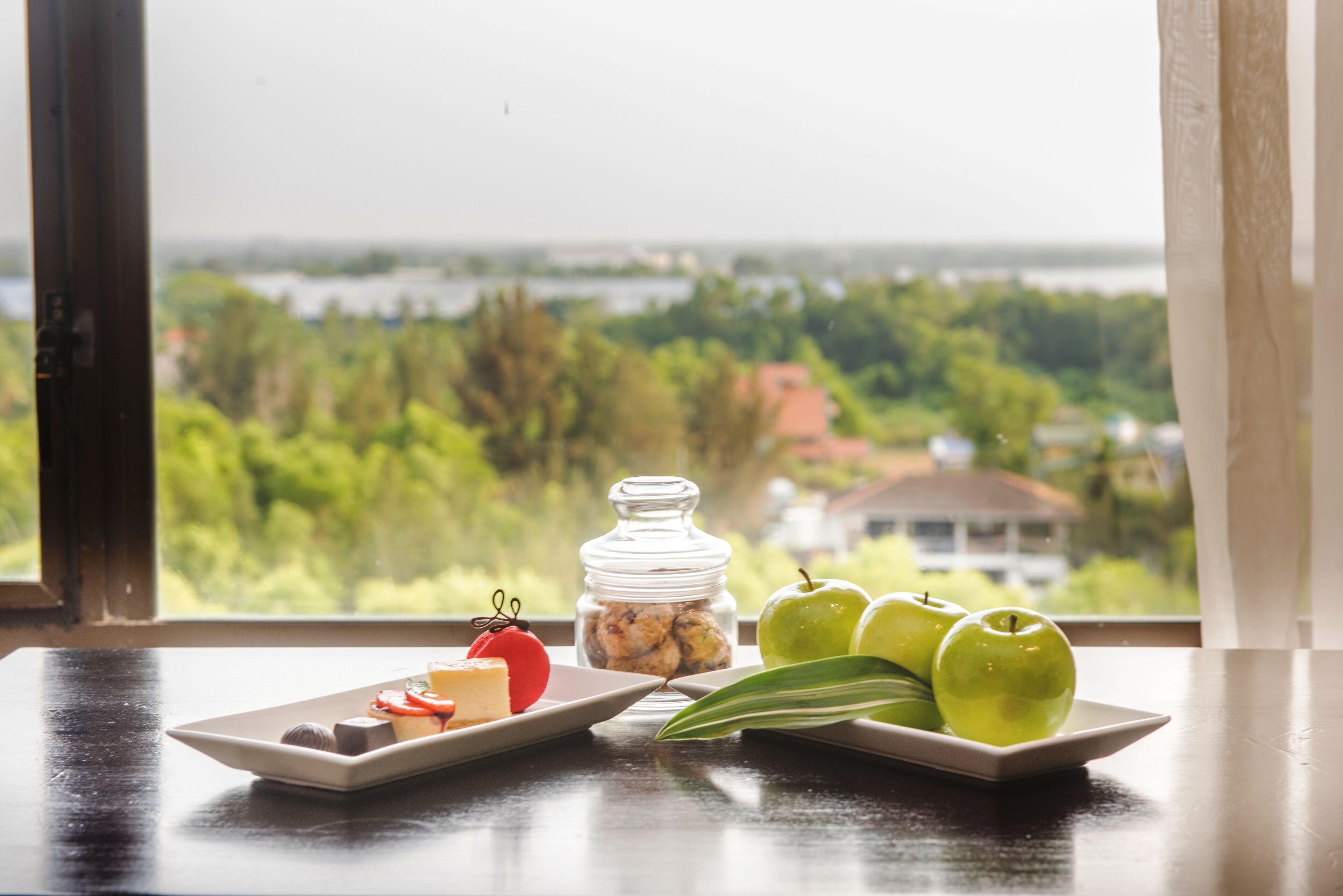 Una mesa con un plato de comida y un tarro de nueces, un plato de manzanas verdes, y una vista por la ventana a un paisaje verde.
