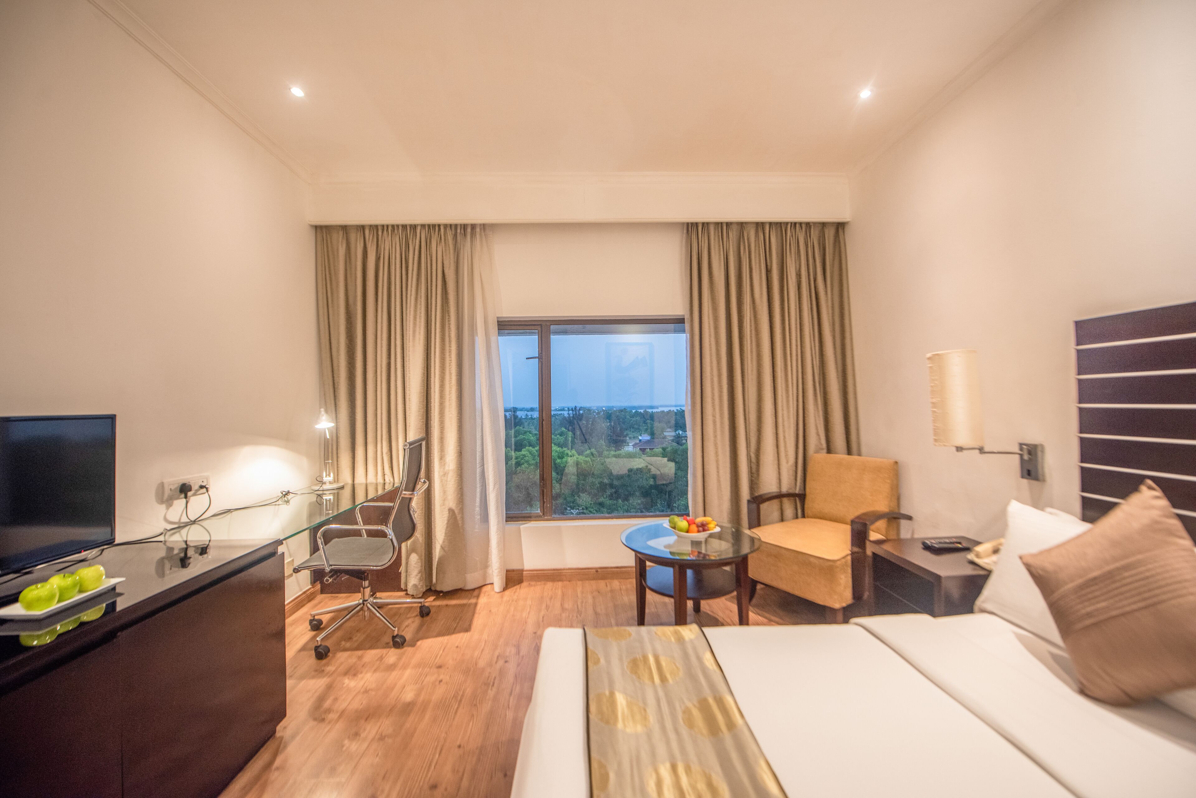 A hotel room with a bed, a desk with a computer, a chair, a small table with fruits, and a TV.