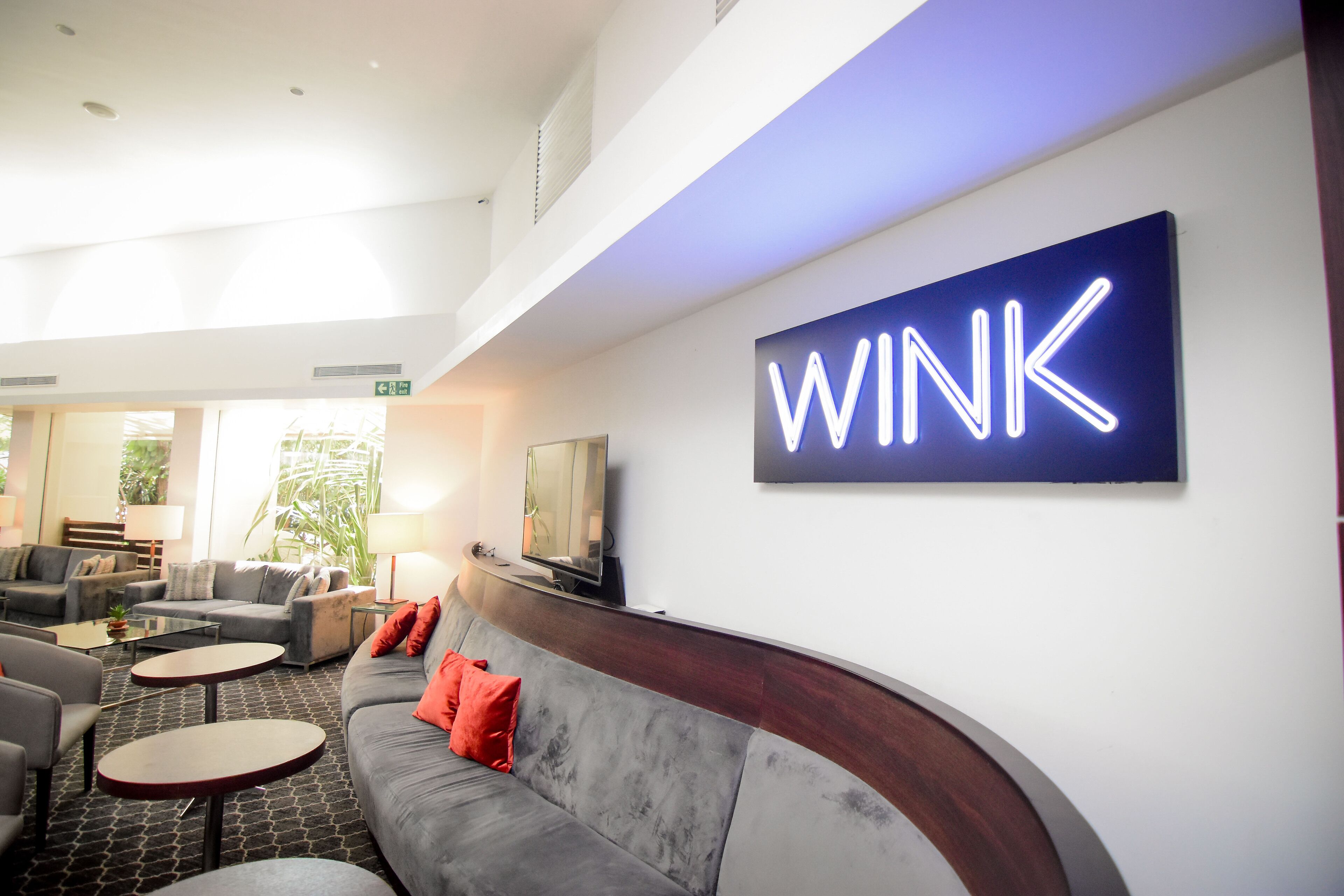 A modern lounge area with a curved grey sofa, red cushions, and a neon sign that reads "WINK".